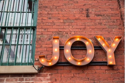 A light-up sign on a brick wall reminding you to cultivate joy.